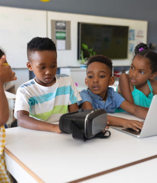 African american elementary school students using vr glasses and laptop at desk in classroom. unaltered, education, virtual reality simulator, wireless technology and school concept. African american elementary school students using vr glasses and laptop at desk in classroom. unaltered, education, virtual reality simulator, wireless technology and school concept.