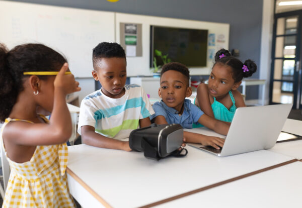 African american elementary school students using vr glasses and laptop at desk in classroom. unaltered, education, virtual reality simulator, wireless technology and school concept.