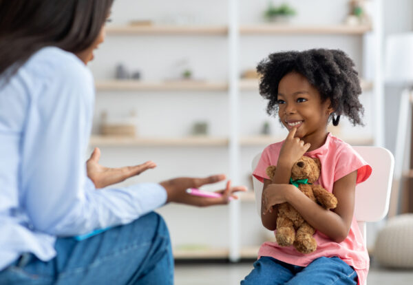 Cheerful cute little african american girl at child psychologists office, sitting on chair in front of unrecognizable female therapist, hugging teddy bear, smiling, feeling safe. Child psychotherapy