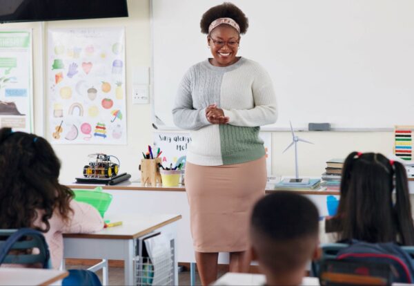 Teacher, children and talking in classroom for lesson, advice or smile with assessment for development. Woman, child and quiz for education, info or knowledge for help, happy or scholarship at school.