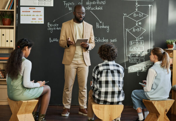 African American teacher using tablet pc and giving lecture to children standing against blackboard at IT lesson