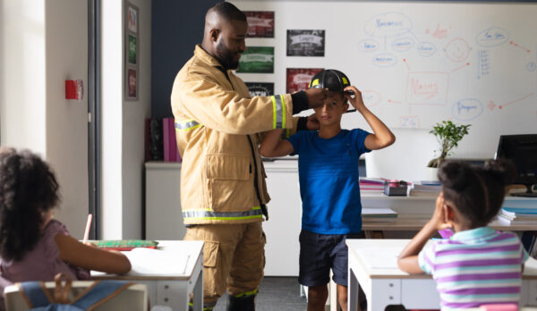 African american young male teacher wearing protective helmet to caucasian elementary boy in class. unaltered, education, firefighter, safety, protection, teaching and school concept.