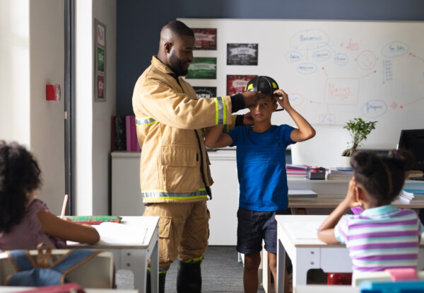 African american young male teacher wearing protective helmet to caucasian elementary boy in class. unaltered, education, firefighter, safety, protection, teaching and school concept.