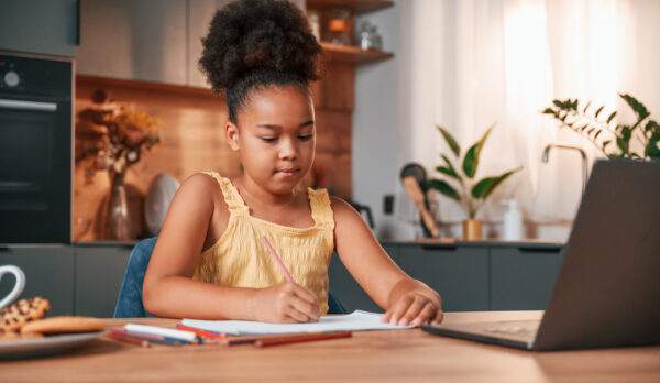 Sitting by the table with laptop and paper for drawing. Cute black girl in casual clothes is at home at daytime Sitting by the table with laptop and paper for drawing. Cute black girl in casual clothes is at home at daytime.