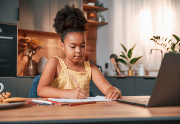 Sitting by the table with laptop and paper for drawing. Cute black girl in casual clothes is at home at daytime.