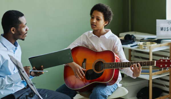 Youthful African American schoolgirl playing acoustic guitar at lesson in music school while sitting in front of teacher consulting her