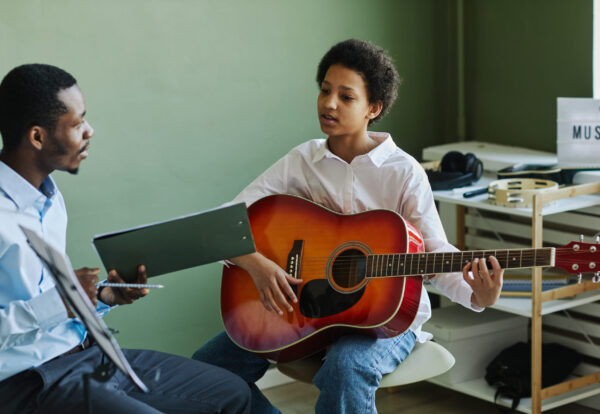 Youthful African American schoolgirl playing acoustic guitar at lesson in music school while sitting in front of teacher consulting her